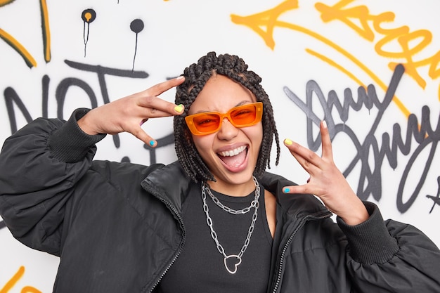 Photo gratuite enthousiaste cool femme ethnique avec des dreadlocks fait un geste de plaisir vêtu d'une veste noire et d'élégantes lunettes de soleil orange sourit largement pose contre le mur de graffitis