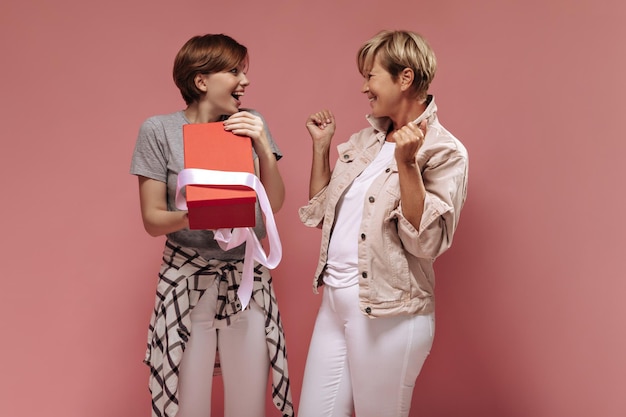 Enthousiaste deux femmes avec une coiffure courte et élégante en pantalon blanc se regardant et posant avec une boîte cadeau rouge ouverte sur fond rose