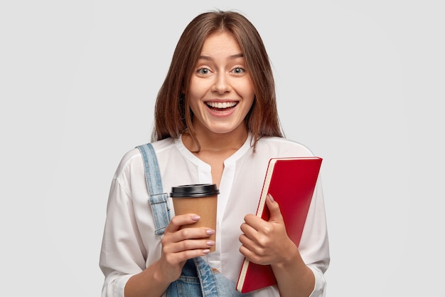 Enthousiaste étudiant diplômé porte une chemise blanche, tient un café et un ordinateur portable à emporter, sourit positivement, isolé sur un mur blanc. Modèles de stagiaires féminins à l'intérieur, satisfaits de quelque chose