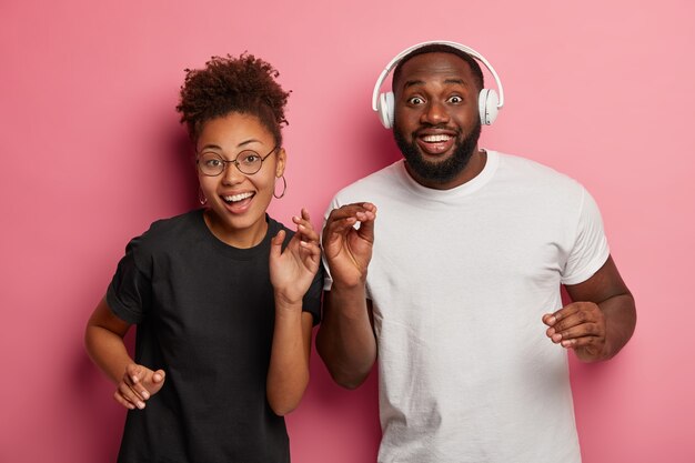 Enthousiaste femme afro à lunettes rondes et son petit ami danse et écoute de la musique, amusez-vous à la fête, de larges sourires, isolés sur un mur rose.