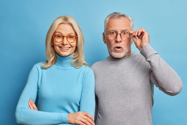 Enthousiaste femme d'âge moyen et son mari retraité réagit sur des nouvelles choquantes garde la main sur des lunettes isolé sur mur bleu