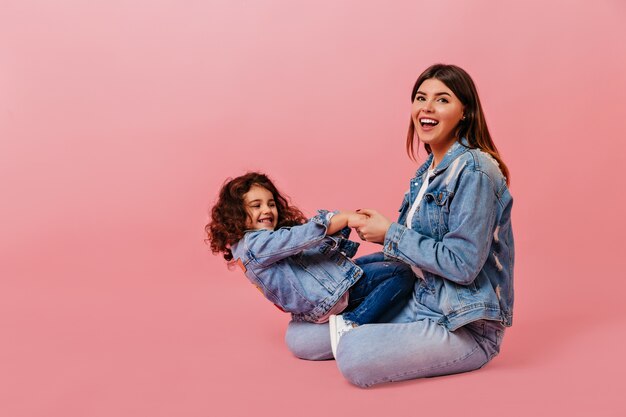 Enthousiaste femme caucasienne jouant avec sa fille. Photo de Studio d'enfant bouclé préadolescent assis avec maman sur fond rose.