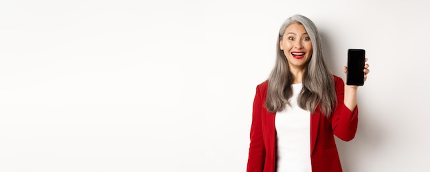 Enthousiaste femme entrepreneur asiatique montrant un écran de téléphone portable souriant amusé debout sur une ba blanche