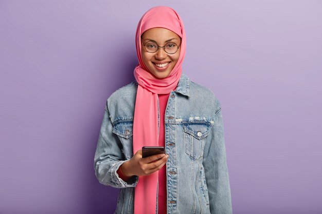 Enthousiaste femme ethnique avec un sourire doux, fait défiler les actualités sur internet sur téléphone mobile, lit un message d'invitation intéressant, habillé en hija rose et lunettes rondes, isolé sur un mur violet