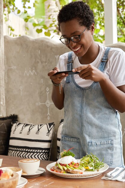 Enthousiaste femme noire fait la photo de la nourriture exotique à la cafétéria, boit du latte