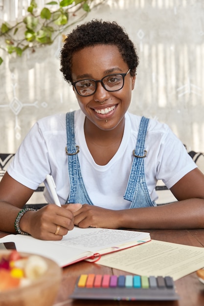 Photo gratuite enthousiaste femme à la peau sombre avec coupe de cheveux afro, apprend le matériel pour l'examen universitaire