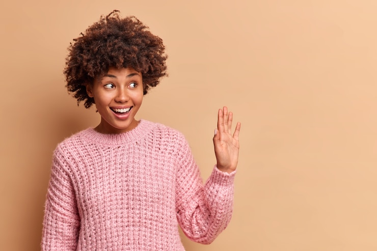 Photo gratuit enthousiaste femme sympathique vagues paume en bonjour geste rencontre quelqu'un à la rue sourit positivement habillé en pull tricoté se dresse à l'intérieur contre un mur marron avec copie espace pour votre information