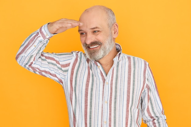 Enthousiaste homme âgé attrayant avec barbe grise et tête chauve posant isolé en gardant la main sur ses yeux pour se protéger