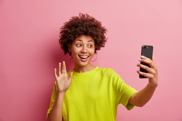 Enthousiaste jeune femme afro-américaine avec des vagues de cheveux cury dans l'appareil photo du smartphone fait un geste salut tout en ayant des entretiens de vidéoconférence avec le meilleur ami à distance porte des t-shirts occasionnels