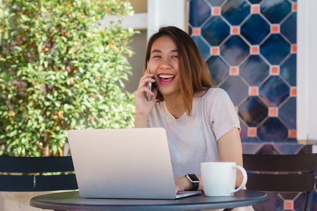 Enthousiaste jeune femme asiatique assise dans un café, boire du café et utiliser un smartphone pour parler