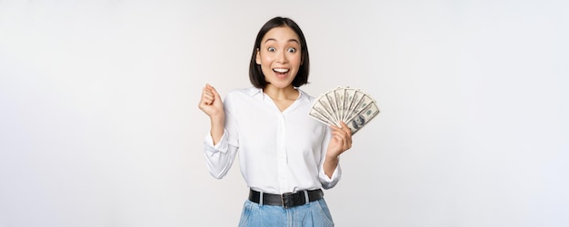 Enthousiaste jeune femme asiatique regardant excitée à la caméra tenant de l'argent en dollars à la main debout sur fond blanc