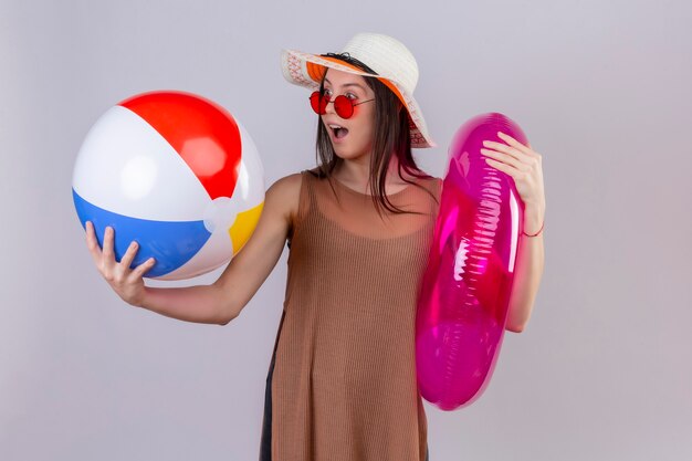 Enthousiaste jeune femme en chapeau d'été portant des lunettes de soleil rouges tenant un anneau gonflable et une balle en regardant surpris et heureux debout