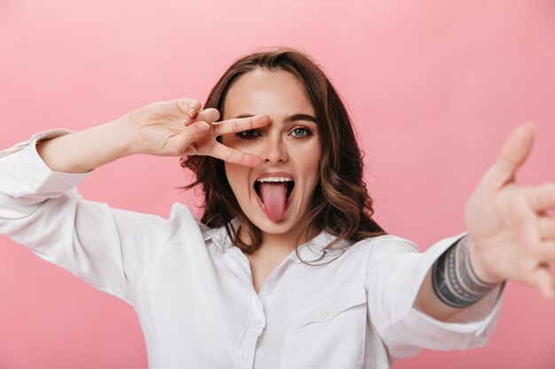 Enthousiaste jeune femme en chemise blanche montre la langue et montre le signe de la paix Brunette fille prend selfie sur fond rose isolé