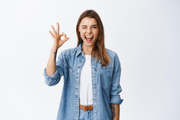 Enthousiaste jeune femme clignant de l'œil et montrant le signe OK dire d'accord ou oui approuver quelque chose de bon recommandant le produit et la qualité debout heureux sur fond blanc