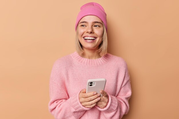 Enthousiaste jeune femme utilise un téléphone portable envoie des messages texte regarde ailleurs a un large sourire commande quelque chose dans la boutique en ligne porte un pull rose et un chapeau isolé sur fond marron Concept technologique