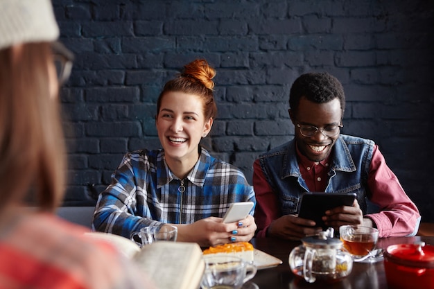 Photo gratuite enthousiaste jeune homme africain portant des lunettes élégantes et jolie femme de race blanche aux cheveux roux ayant une conversation de souris avec une fille brune méconnaissable devant eux