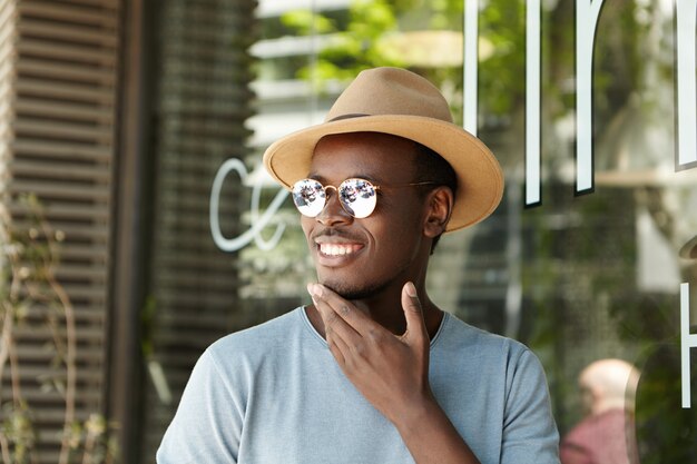 Enthousiaste jeune homme afro-américain en lunettes de soleil à la mode et couvre-chef touchant son menton et souriant joyeusement alors qu'il voit son ami s'approcher de lui en attendant le déjeuner au restaurant sur le trottoir