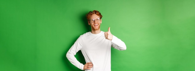 Enthousiaste jeune homme à la barbe et aux cheveux rouges portant des lunettes montrant le pouce en signe d'approbation et souriant s