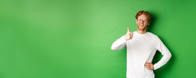 Photo gratuite enthousiaste jeune homme à la barbe et aux cheveux rouges portant des lunettes montrant le pouce en signe d'approbation et souriant s