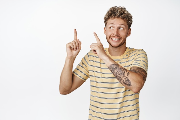 Enthousiaste jeune homme blond montrant une publicité pointant du doigt le coin supérieur gauche et souriant excité recommandant la vente en magasin fond blanc