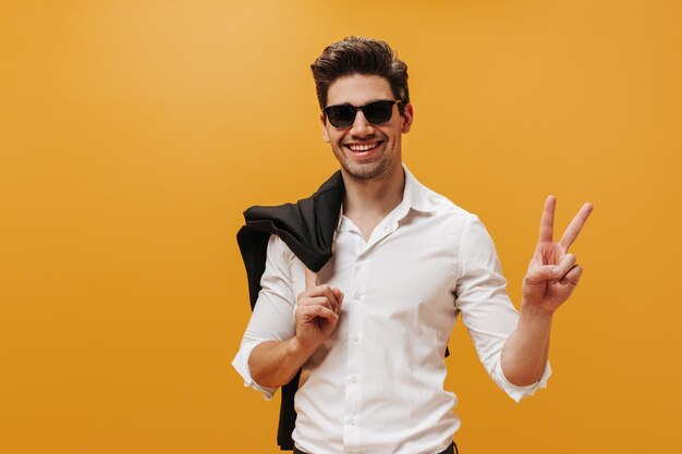 Enthousiaste jeune homme brun en lunettes de soleil et chemise blanche montre un signe de paix et tient une veste sur fond orange isolé