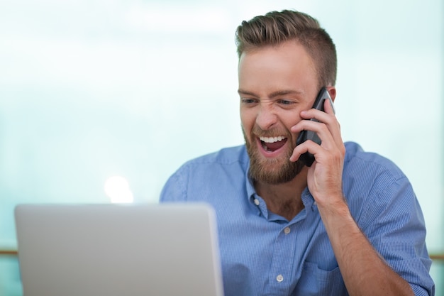 Enthousiaste jeune homme parlant au téléphone avec un ordinateur portable