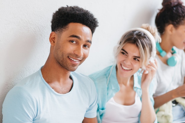 Enthousiaste jeune homme à la peau brune et grand sourire posant à côté du mur blanc. Portrait d'heureux mec aux cheveux noirs et fille riante dans de grands écouteurs appréciant la musique.