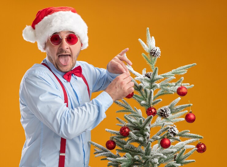 Photo gratuit enthousiaste jeune homme portant des bretelles noeud papillon en bonnet de noel et lunettes rouges debout près de l'arbre de noël jouet suspendu sur l'arbre heureux et surpris qui sort la langue sur fond orange