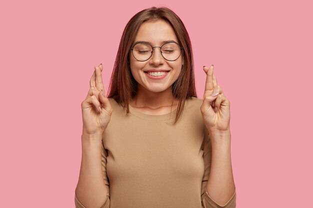 Enthousiaste jeune jolie jeune femme avec une expression positive croise les doigts, espère la réalisation complète du rêve, fait des vœux, porte des lunettes rondes et un pull décontracté, isolé sur un mur rose.