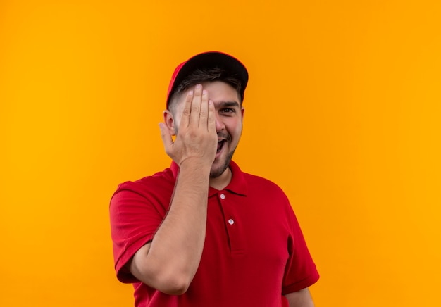 Enthousiaste jeune livreur en uniforme rouge et capuchon fermant un œil avec une main souriant largement