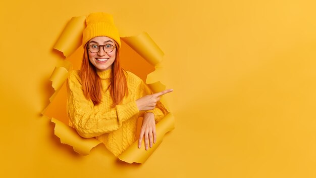 Enthousiaste jeune mannequin a les cheveux roux à pleines dents sourire pointant vers l'espace de copie, vêtue de lunettes de pull jaune.