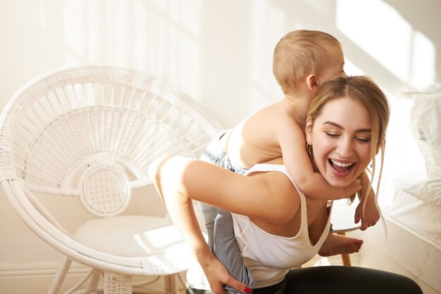 Enthousiaste jeune mère s'amusant à l'intérieur donnant ferroutage à son mignon petit fils. Jolie maman passant du temps à la crèche, jouant avec l'enfant pendant la distance sociale à cause de la quarantaine