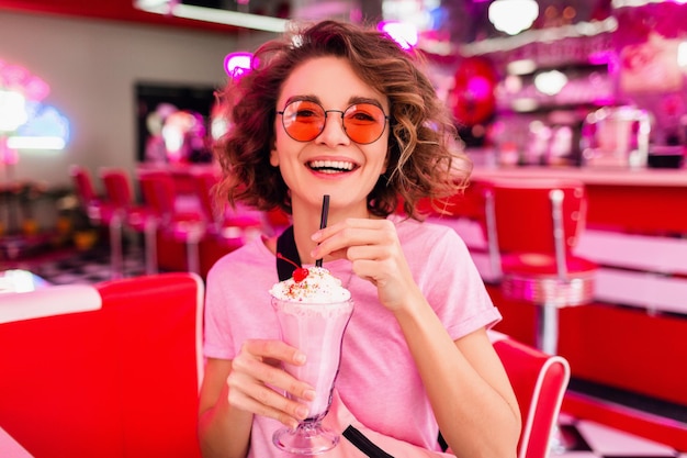 Enthousiaste jolie femme souriante élégante dans un café rétro vintage des années 50 assis à table en train de boire un cocktail de milk-shake portant des lunettes de soleil roses s'amusant à rire avec un sourire candide