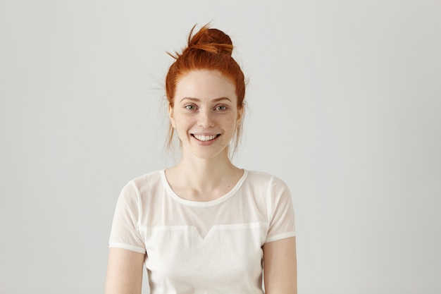 Enthousiaste magnifique jeune femme portant ses cheveux roux en noeud souriant joyeusement tout en recevant des nouvelles positives. Jolie fille vêtue d'un chemisier blanc à la recherche d'un sourire joyeux excité