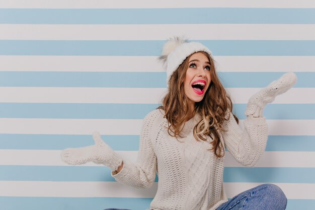 Enthousiaste modèle jeune fille bouclée en vêtements blancs et blue-jeans est assis sur le sol et posant émotionnellement pour le portrait sur le mur bleu rayé