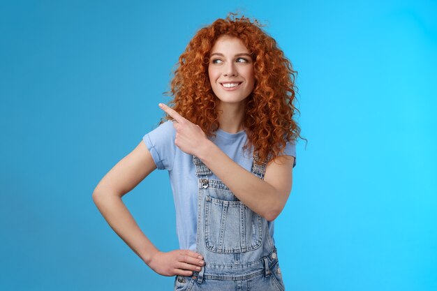 Enthousiaste motivé charismatique beau heureux rousse souriante 25s femme aux cheveux bouclés salopette d'été pointant vers le coin supérieur gauche regarder de côté heureux montrer la promo suggérer l'essai. Espace de copie