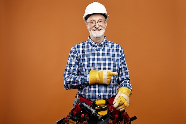 Enthousiaste à la recherche frinedly caucasian senior male menuisier pointant l'index portant l'équipement de travail autour de la taille. Électricien barbu âgé souriant posant dans des gants de protection et une ceinture à outils