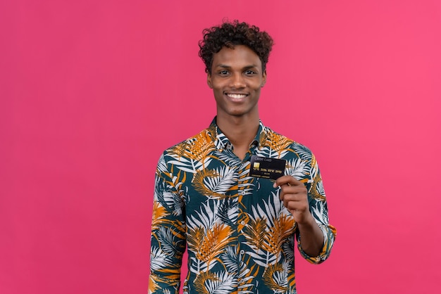 Enthousiaste et souriant jeune bel homme à la peau foncée avec des cheveux bouclés en chemise imprimée de feuilles tout en tenant et en montrant la carte de crédit