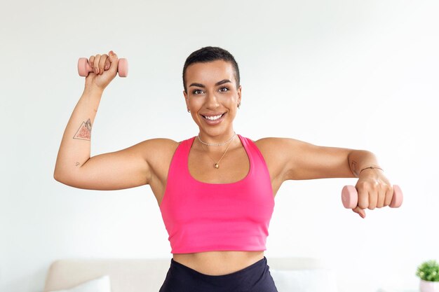 Entraînement domestique avec des poids Dame noire positive faisant des exercices avec des haltères renforçant son corps à la maison Souriante jeune femme travaillant sur ses muscles du biceps en restant en bonne santé