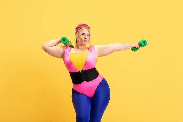 Entraînement d'un jeune modèle féminin de taille plus caucasienne sur un mur jaune. Espace de copie. Concept de sport, mode de vie sain, corps positif, mode, style. Femme élégante pratiquant avec des poids verts.