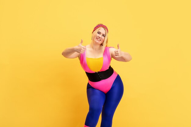 Entraînement d'un jeune modèle féminin de taille plus caucasienne sur un mur jaune. Espace de copie. Concept de sport, mode de vie sain, corps positif, mode, style. Femme élégante souriante avec les pouces vers le haut.