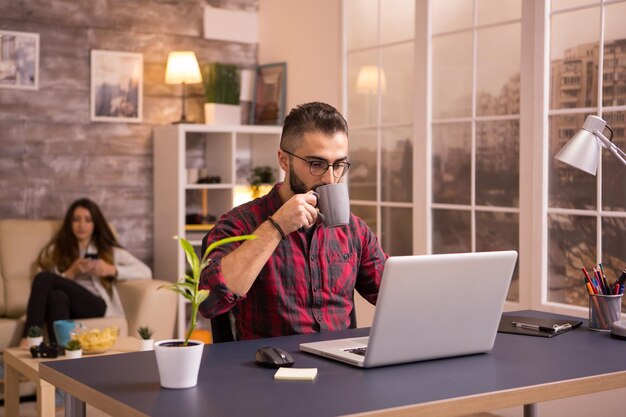 Entrepreneur caucasien barbu prenant une gorgée de café tout en travaillant sur un ordinateur portable dans le salon. Petite amie sur le canapé en arrière-plan parcourant le téléphone. Jetons sur table.