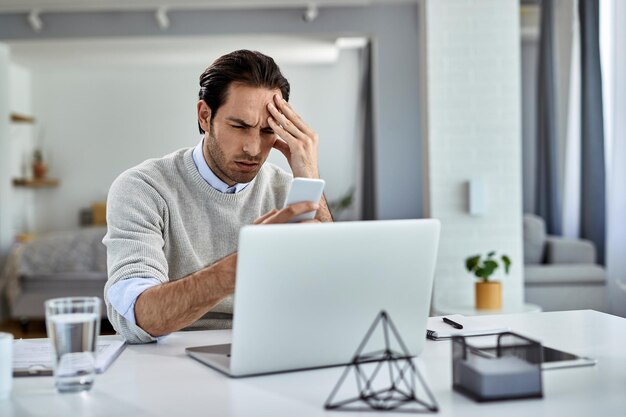 Entrepreneur désemparé travaillant à la maison et tenant sa tête dans la douleur tout en lisant un message texte sur un téléphone portable