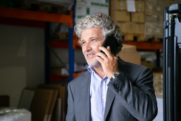 Entrepreneur Mature Positif Pensif Debout Dans L'entrepôt Et Parlant Au Téléphone Mobile. étagères Avec Des Marchandises En Arrière-plan. Copiez L'espace. Concept D'entreprise Ou De Communication