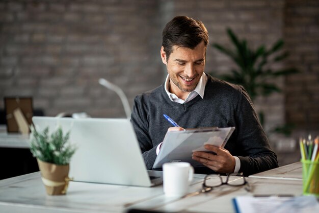 Entrepreneur souriant écrivant un rapport d'activité tout en travaillant sur un ordinateur portable au bureau