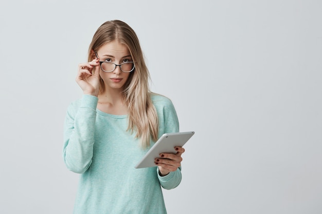 Entrepreneuse blonde confiante dans des lunettes élégantes avec tablette contre un mur gris, travaille au développement d'un nouveau projet. Jeune enseignant à lunettes utilise la technologie moderne