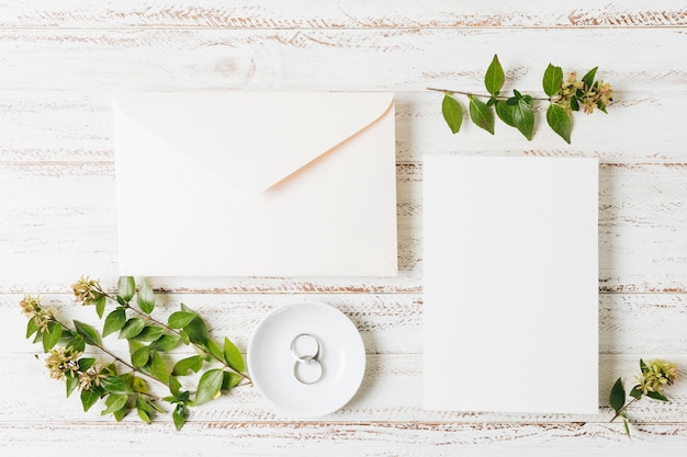 Enveloppe fermée; carte; rameau de fleurs et alliances sur une assiette au-dessus du bureau blanc