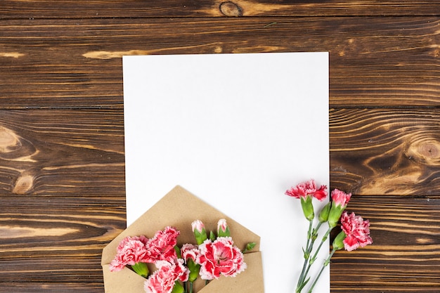 Enveloppe avec des fleurs d&#39;oeillets rouges et du papier blanc vierge sur une surface en bois
