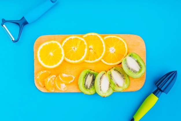 Photo gratuite Éplucheur et presse-agrumes à la main avec des oranges et des tranches de kiwi sur une planche à découper sur fond bleu