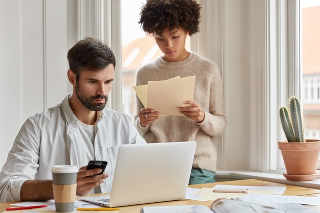 Une équipe diversifiée d'employés a des réunions informelles dans une atmosphère domestique, travaille avec des documents papier, développe le démarrage pendant le briefing. Le PDG masculin sérieux concentré détient les chèques de téléphone portable sur un ordinateur portable